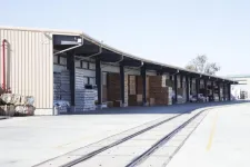Thumbnail for Outdoor warehouse with stacked boxes and pallets under a long covered loading dock near railway tracks.