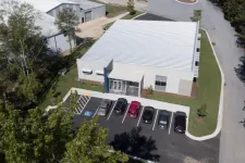 Thumbnail for Aerial view of a modern commercial building with a parking lot and six parked cars surrounded by trees and a road.
