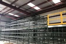 Thumbnail for Empty metal storage racks inside a warehouse with a yellow safety gate on the upper platform under a skylit roof.