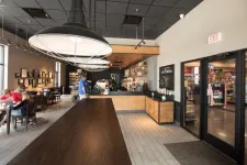 Thumbnail for Modern coffee shop interior with wooden tables, large pendant lights, and customers sitting near windows.