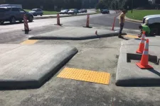 Thumbnail for Newly constructed concrete curb ramps with tactile paving and traffic cones near a busy road under clear sky.
