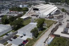 Thumbnail for Aerial view of an industrial facility with warehouses, parked cars, stacks of metal beams, and railway tracks.
