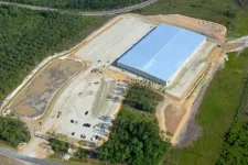 Thumbnail for Aerial view of a large warehouse under construction surrounded by cleared land and forested areas.