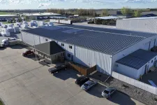 Thumbnail for Aerial view of a large industrial warehouse with parked cars and stacks of materials outside under a partly cloudy sky
