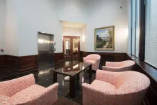 Thumbnail for Modern lobby with pink patterned armchairs, glass coffee table, elevator, and large windows with blinds.