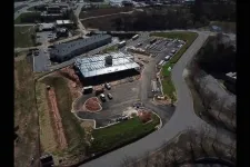 Thumbnail for Aerial view of a construction site with a building framework, surrounding roads, parking, and vehicles during daytime.