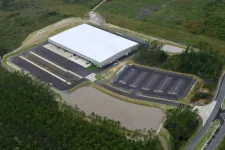 Thumbnail for Aerial view of a large white-roof warehouse surrounded by parking lots, a pond, and green wooded areas.