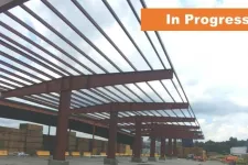 Thumbnail for Steel frame construction of a large building in progress under a cloudy sky with stacked wood and equipment nearby