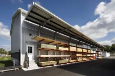 Thumbnail for Outdoor lumber storage racks under metal roof with various wood planks on a sunny day with blue sky and clouds.