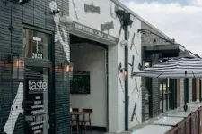 Thumbnail for Exterior view of Taste Wine Bar and Market with black and white walls and outdoor seating under umbrella.