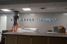 Thumbnail for Worker on ladder installing illuminated sign that reads Savor Repeat on tiled wall in modern interior.