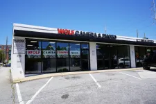 Thumbnail for Exterior of Wolf Camera & Photo store with signage and parking lot on a clear sunny day.
