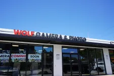 Thumbnail for Storefront of Wolf Camera & Photo with signage and Coming Soon banners under a clear blue sky.