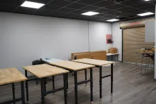 Thumbnail for Empty workshop room with wooden tables, black chairs, gray walls, and a roll-up door on wood flooring.