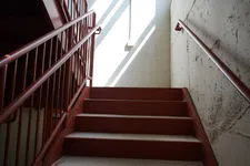 Thumbnail for Red metal staircase with handrails leading up to a sunlit white brick wall and window.