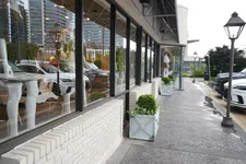 Thumbnail for Sidewalk view of a modern store with large windows, potted plants, street lamps, and parked cars on a rainy day.