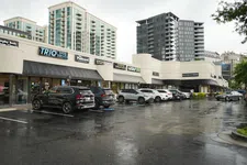 Thumbnail for Shopping center with parked cars on wet pavement under cloudy sky in urban area with high-rise buildings.