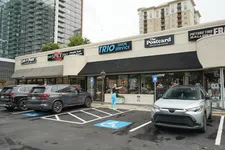 Thumbnail for Street view of small retail shops including Trio Shoe Service, The Postcard cafe, and parking with cars and a person in bright pants.