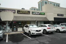 Thumbnail for Strip mall storefronts with shops named Cookie Creations, GolfTec, and Moda 404 with cars parked in front on a cloudy day