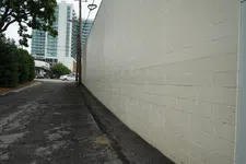 Thumbnail for Empty urban alley with cracked pavement, beige brick wall, trees, and distant modern buildings under cloudy sky