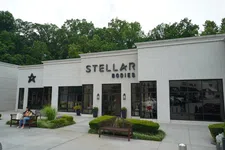 Thumbnail for Exterior of Stellar Bodies store with large windows, entrances, benches, and greenery on a bright day.