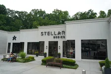 Thumbnail for Modern white commercial building with Stellar Bodies signage, large windows, benches, greenery, and a person sitting outside.