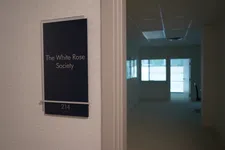 Thumbnail for Office door sign for The White Rose Society, room 214, with a dimly lit hallway and office visible inside.