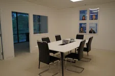 Thumbnail for Modern conference room with white table, six black chairs, water bottles, and framed accolades on wall.