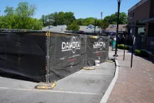 Thumbnail for Outdoor construction site fenced with black Dakota-branded mesh screens along a brick sidewalk under clear sky.
