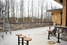 Thumbnail for Outdoor patio with stone wall, wooden stools, metal ladder, and worker using tools near building corner