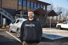Thumbnail for Smiling man in glasses wearing a black Dakota hoodie stands in a parking lot with cars and a modern building in the background.