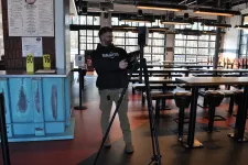 Thumbnail for Man adjusting camera on tripod inside a modern cafe with empty tables and bar stools during daytime.