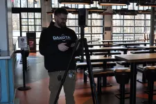 Thumbnail for Man adjusting camera equipment on tripod inside a modern empty restaurant with large windows and tables.