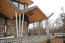 Thumbnail for Modern building exterior with angled wooden roof, white pillars, stone wall, and outdoor ceiling fans overlooking a garden.
