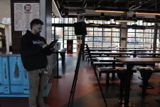 Thumbnail for Man wearing Dakota sweatshirt using tablet near a tripod in a spacious, modern restaurant with large windows.
