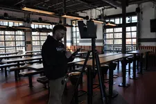 Thumbnail for Man in a black hoodie using a tablet next to a speaker on a tripod inside a modern empty restaurant with wooden tables.