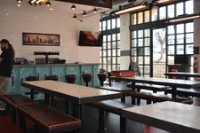 Thumbnail for Modern indoor bar with long tables, leather benches, bar stools, large windows, and industrial lighting fixtures.