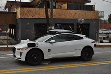 Thumbnail for White Waymo autonomous electric vehicle with sensors parked on city street in front of modern building.