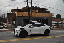 Thumbnail for White Waymo self-driving electric car parked on a city street with modern building in the background.