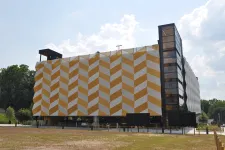 Thumbnail for Modern building with a yellow and white geometric facade and glass elevator tower under a partly cloudy sky.