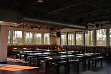 Thumbnail for Empty modern cafeteria with large windows, wooden benches, and overhead ventilation ducts during daytime.