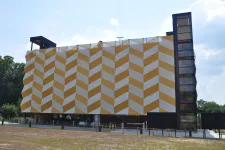 Thumbnail for Modern building with yellow and white geometric patterned facade and glass elevator tower on a sunny day.