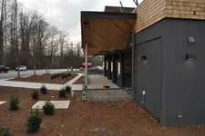 Thumbnail for Modern building exterior with wooden and black brick facade beside a bare landscaped area and road with vehicles.