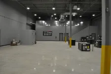 Thumbnail for Spacious industrial warehouse interior with gray walls, concrete floor, HVAC units, and bright overhead lights.