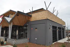 Thumbnail for Modern restaurant exterior with gray brick, large windows, and angled wooden roof under cloudy sky.