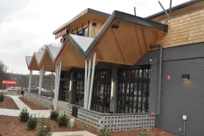 Thumbnail for Modern building exterior with angled wooden roof panels, large glass windows, and landscaped surroundings on a cloudy day