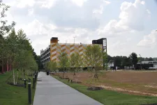 Thumbnail for Pathway lined with young trees and lights next to a yellow and white patterned building under a cloudy sky.