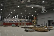 Thumbnail for Large industrial warehouse under construction with workers, scaffolding, pallets, and HVAC ducts visible.