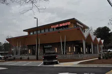 Thumbnail for Modern Killer Burgers Grindhouse restaurant with distinctive angular roof and parking lot under cloudy sky.