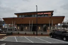 Thumbnail for Modern Killer Burgers restaurant exterior with large wood awning and parking spaces under cloudy sky